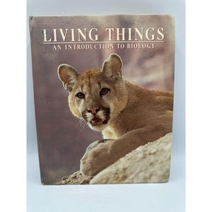 May include: A vintage book titled "LIVING THINGS AN INTRODUCTION TO BIOLOGY" features a cougar on the cover. The cougar is tan with dark eyes and is resting on a rock. The book has a brown and tan color scheme.