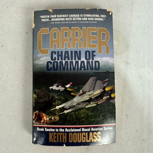 Peut inclure: Un livre de poche intitulé "Carrier: Chain of Command" de Keith Douglass. La couverture présente des avions de chasse en vol au-dessus de l'eau, avec le mot "Carrier" en grandes lettres dorées. Le livre fait partie de la "Acclaimed Naval Aviation Series".