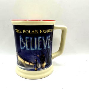 May include: Cream-colored ceramic mug with a red rim and a large handle. The mug features a night scene with the words "THE POLAR EXPRESS BELIEVE" in a blue font. A boy and a man are standing in the snow.