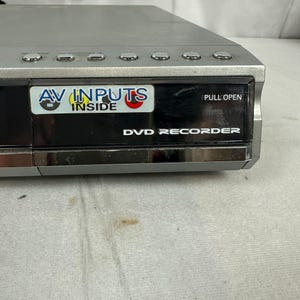 Pode incluir: Gravador de DVD prateado com um painel frontal preto. O painel tem o texto "AV INPUTS INSIDE" e "DVD RECORDER". O dispositivo tem v&aacute;rios bot&otilde;es na parte superior e uma etiqueta "PULL OPEN".