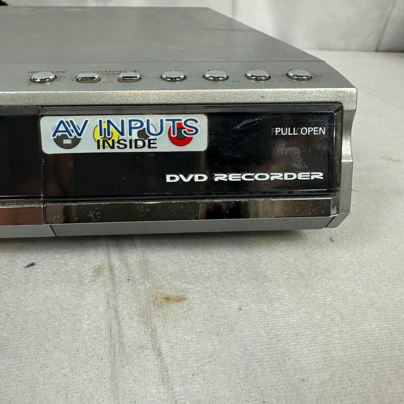 Pode incluir: Gravador de DVD prateado com um painel frontal preto. O painel tem o texto "AV INPUTS INSIDE" e "DVD RECORDER". O dispositivo tem v&aacute;rios bot&otilde;es na parte superior e uma etiqueta "PULL OPEN".