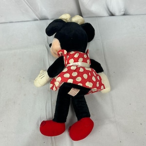 May include: A Minnie Mouse soft toy, seen from behind, with a black body, a red and white polka dot dress, and red shoes. The doll has white gloves and a cream-coloured bow on her head. A classic Disney character.