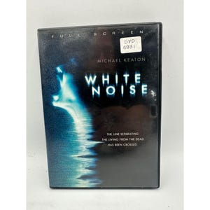 Peut inclure: Un boîtier DVD du film "White Noise" avec Michael Keaton. La couverture présente un fond sombre avec une image blanche et bleue d'un visage hurlant et le titre du film. Le texte "La ligne séparant les vivants des morts a été franchie" est également visible.