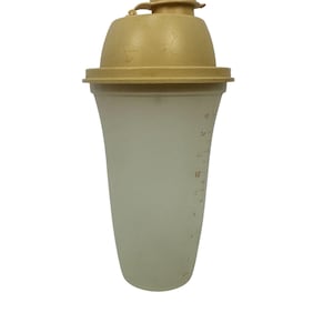 Puede incluir: Un agitador de plástico blanco roto vintage con tapa y tapón color beige. El agitador tiene marcas de medición en onzas líquidas en el lateral. La tapa es redonda y tiene un pequeño tapón abatible. El agitador tiene forma acampanada.