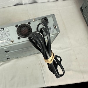 Pode incluir: Um gravador de DVD prateado com um cabo de alimenta&ccedil;&atilde;o preto preso com uma faixa el&aacute;stica bege. A parte traseira do dispositivo possui uma ventoinha, etiquetas de aviso e uma certifica&ccedil;&atilde;o UL.