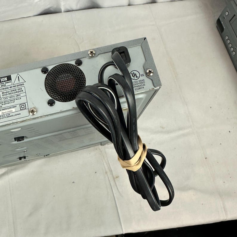 Pode incluir: Um gravador de DVD prateado com um cabo de alimenta&ccedil;&atilde;o preto preso com uma faixa el&aacute;stica bege. A parte traseira do dispositivo possui uma ventoinha, etiquetas de aviso e uma certifica&ccedil;&atilde;o UL.