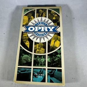 Pode incluir: Uma fita VHS vintage para "Family Opry Reunion Volume Five". A capa apresenta uma colagem de artistas em um padrão de grade, com o logotipo Opry em azul e branco. A fita tem um esquema de cores verde claro e bege.
