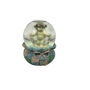 May include: A snow globe featuring a cherubic angel with a floral crown and garland. The angel stands inside a clear glass globe atop a patchwork-style base with a tied bow. The base is primarily blue and green.