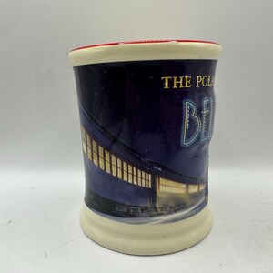 May include: A ceramic mug with a cream-colored base and rim, and a red interior rim. The mug features a night scene with a train and the words "THE POLAR BE". The background is dark blue.