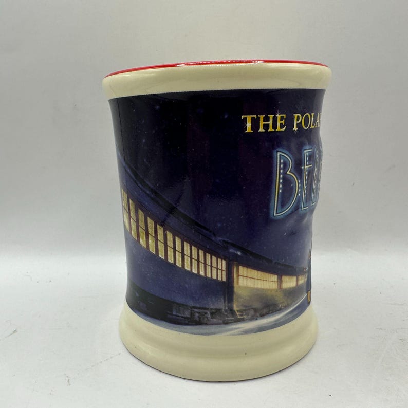 May include: A ceramic mug with a cream-colored base and rim, and a red interior rim. The mug features a night scene with a train and the words "THE POLAR BE". The background is dark blue.