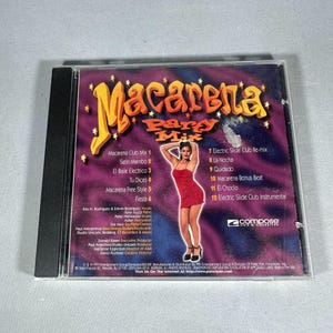 Puede incluir: Una caja de CD con el título "Macarena Party Mix" en letras amarillas y naranjas. La portada presenta a una mujer con un vestido rojo y una lista de canciones. La caja del CD está en una funda de plástico negro.