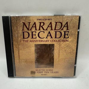 Op de afbeelding: Een dubbel-cd-set getiteld Narada Decade: The Anniversary Collection, Selected Works The First Ten Years. De hoes toont een goudbruin landschap met bomen en een centrale rechthoekige afbeelding van een bos, verpakt in een doorzichtig plastic doosje.
