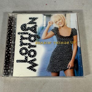 Peut inclure: Un boîtier de CD avec l'album "Shakin' Things Up" de Lorrie Morgan. La pochette montre une femme dans une robe à pois noirs et blancs. Le titre de l'album est en lettres blanches à pois.