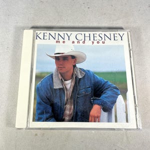Puede incluir: Una caja de CD con el álbum "me and you" de Kenny Chesney. La portada muestra a un hombre con sombrero de vaquero y chaqueta vaquera apoyado en una valla blanca. El título del álbum y el nombre del artista están impresos en blanco.