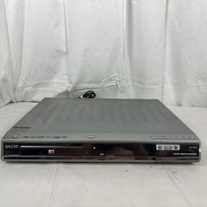 Pode incluir: Um gravador de DVD Sanyo prateado com um painel frontal preto. O dispositivo possui uma bandeja de disco, v&aacute;rios bot&otilde;es e um cabo de alimenta&ccedil;&atilde;o. O painel frontal tem o texto "DVD Recorder" e o logotipo da Sanyo.