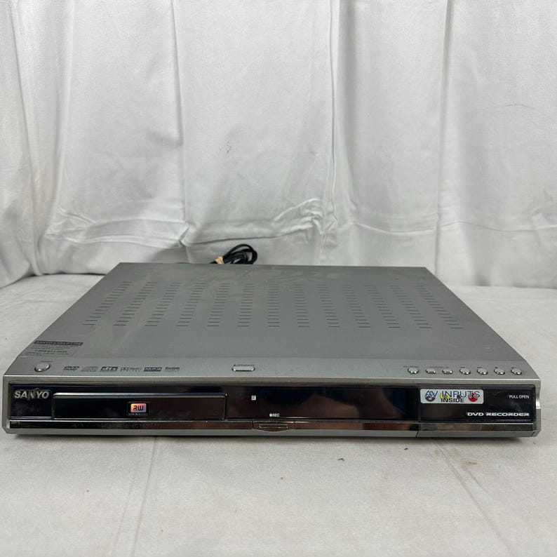 Pode incluir: Um gravador de DVD Sanyo prateado com um painel frontal preto. O dispositivo possui uma bandeja de disco, v&aacute;rios bot&otilde;es e um cabo de alimenta&ccedil;&atilde;o. O painel frontal tem o texto "DVD Recorder" e o logotipo da Sanyo.