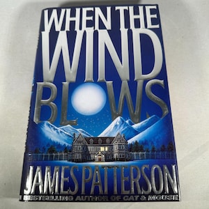 May include: A hardcover book titled "When the Wind Blows" by James Patterson. The cover features a nighttime scene with a full moon, mountains, and a large house. The title and author's name are in silver lettering.