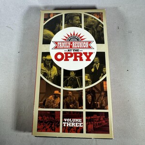 Pode incluir: Uma caixa retangular de fita VHS com uma colagem de imagens a preto e branco de artistas. A caixa apresenta o texto "COUNTRY'S FAMILY-REUNION AT THE OPRY" e "VOLUME THREE" em vermelho e branco.