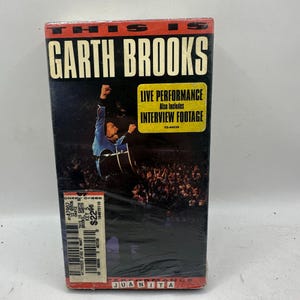 Pode incluir: Uma fita VHS com Garth Brooks. A capa apresenta Brooks atuando ao vivo, com o texto "LIVE PERFORMANCE" e "INTERVIEW FOOTAGE". A fita está em uma embalagem plástica.
