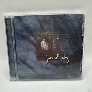 May include: A CD case featuring the album "Jars of Clay." The cover art displays a dark, textured background with a faded image of four people. The band's name is written in a cursive font.