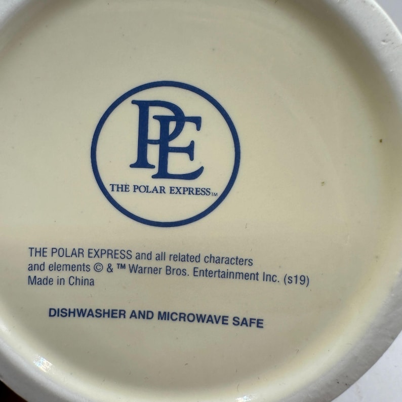 May include: Cream-colored ceramic mug with a blue circular logo featuring the letters "P" and "E" and the text "THE POLAR EXPRESS". The text "DISHWASHER AND MICROWAVE SAFE" is also visible.