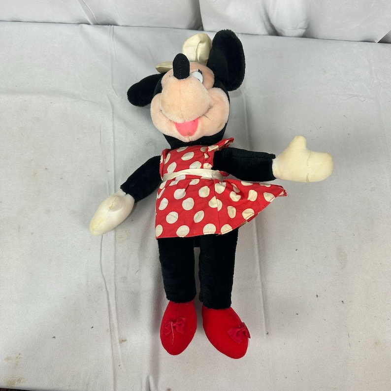 May include: A Minnie Mouse soft toy. The toy has black ears, arms, and legs, a pink face, and a red dress with white polka dots. The toy is wearing red shoes and a white bow on her head.