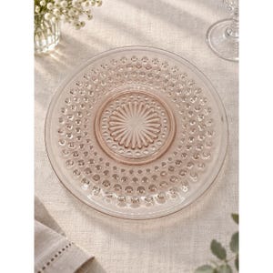 May include: A round, pale pink glass plate featuring a textured surface with a repeating pattern of raised dots along the rim and a central sunburst design consisting of radiating lines. The plate has a smooth, translucent finish.