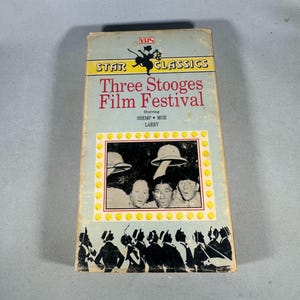 Könnte beinhalten: Eine Vintage-VHS-Kassette für das "Three Stooges Film Festival" von Star Classics. Die Kassette hat ein hellblaues Cover mit dem Titel in Rot und den Gesichtern der Stooges in einem gerahmten Bild. Ein Retro-Sammlerstück.