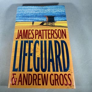 Könnte beinhalten: Buchcover von "LIFEGUARD" von James Patterson & Andrew Gross. Das Bild zeigt eine Strandszene mit einem Rettungsschwimmerstand, einer Person und blauen Meereswellen. Der Titel ist in großen blauen Buchstaben gehalten.