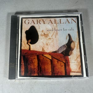 May include: A CD case featuring the album cover for Gary Allan's "Used Heart for Sale." The artwork depicts a person's legs and a hat resting on a wooden surface, with the album title and artist's name prominently displayed.