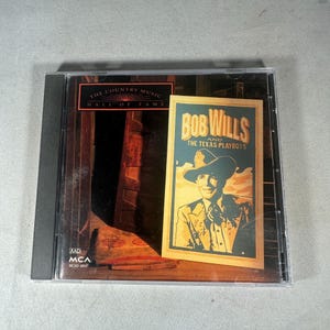 May include: A CD case featuring the album "Bob Wills and The Texas Playboys" from The Country Music Hall of Fame. The album cover has a vintage design with a portrait of Bob Wills in a cowboy hat. The case is in a clear plastic sleeve.