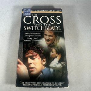 May include: A VHS tape for "The Cross and the Switchblade" with a black cover. The title is in large white letters. The cover features images of people and text that reads "David Wilkerson's Courageous Mission. Nicky Cruz's Dramatic Conversion."