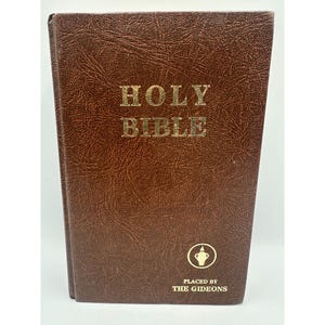 May include: A brown leather-bound Holy Bible with gold lettering. The cover features the words "HOLY BIBLE" in a bold, serif font. Below the title is a small emblem and the text "PLACED BY THE GIDEONS". The book has a textured appearance.