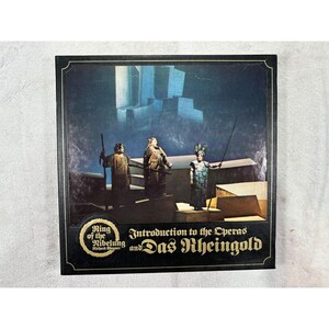 May include: A vintage album cover featuring a stage scene from an opera. The cover has a black border and gold text that reads "Ring of the Nibelung" and "Introduction to the Operas and Das Rheingold". The image shows three costumed figures on a stage.