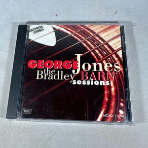 May include: A compact disc case featuring the album "George Jones the Bradley Barn Sessions." The cover has a dark red and black background with the album title in red and white text. A white guitar pick is in the upper left corner.
