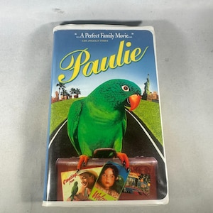 May include: VHS tape for the movie "Paulie". The cover features a vibrant green parrot with an orange beak, standing on a suitcase. The background shows a road, palm trees, and the Statue of Liberty. Text includes "...A Perfect Family Movie..." and "Paulie".