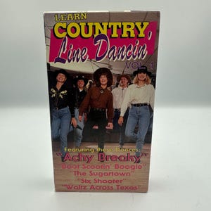 May include: A VHS tape titled "Learn Country Line Dancin' Vol. 1." The cover features a group of people in cowboy hats and jeans. The title is in pink and yellow. The tape lists dances like "Achy Breaky" and "Boot Scootin' Boogie."