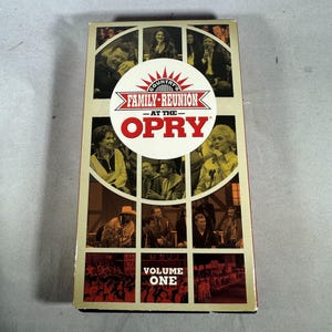 Pode incluir: Uma fita VHS vintage de "Country's Family Reunion at the Opry", Volume One. A capa apresenta uma colagem de artistas e o título em vermelho e branco. A fita está em uma caixa retangular com um esquema de cores bege e marrom.