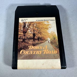 Könnte beinhalten: Schwarzes 8-Spur-Tonband mit einem Cover, das eine von Bäumen in Herbstfarben gesäumte Straße zeigt. Der Text "Down A Country Road" ist in einer stilisierten Schriftart gehalten. Das Band ist mit "A Columbia Musical Treasury" beschriftet.