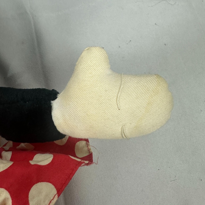 May include: Close-up of a vintage stuffed toy hand. The hand is made of off-white fabric with visible stitching. A black fabric section and a red fabric with white polka dots are also visible.