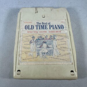 Op de afbeelding: Off-white 8-track tape cartridge met een papieren label. Het label leest "The Best of Old Time Piano" en bevat illustraties van muzikanten. Muziekstijlen zijn Honky Tonk, Ragtime en Barrel House.