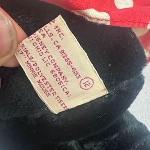 May include: A close-up of a black plush toy with a tag. The tag has text that includes "Disney Company" and "Minnie Mouse". The toy has a red and white polka dot bow.