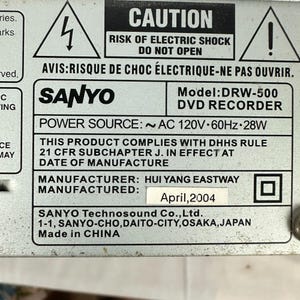 Pode incluir: Close-up de uma placa de metal com texto. A placa tem um aviso sobre choque el&eacute;trico e o texto "SANYO DVD RECORDER". Tamb&eacute;m indica que o produto foi fabricado em abril de 2004.