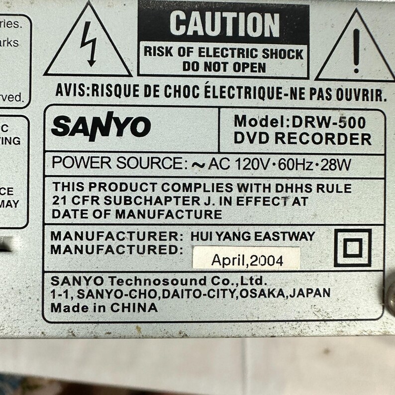 Pode incluir: Close-up de uma placa de metal com texto. A placa tem um aviso sobre choque el&eacute;trico e o texto "SANYO DVD RECORDER". Tamb&eacute;m indica que o produto foi fabricado em abril de 2004.