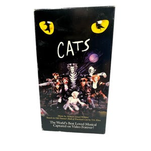 Peut inclure: Boîtier VHS noir du spectacle musical "Cats". La couverture présente un groupe d'artistes costumés avec du maquillage de chat sur fond sombre. Le titre "CATS" est en blanc, avec des yeux de chat jaunes au-dessus. Texte : "Musique d'Andrew Lloyd Webber."