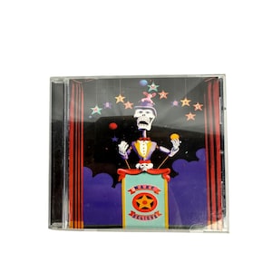 Puede incluir: Una caja de CD con un esqueleto con esmoquin haciendo malabarismos con bolas de colores en un escenario. El escenario tiene cortinas rojas y una pancarta que dice "MAKE BELIEVE" con un emblema de estrella. El fondo es oscuro con luces en forma de estrella.