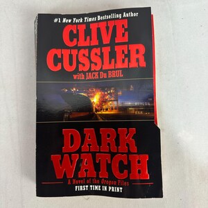 Może przedstawiać: Twarda oprawa książki zatytułowanej "Dark Watch" autorstwa Clive Cusslera i Jacka Du Brula. Okładka jest czarna z czerwonymi literami, prezentującymi tytuł i nazwiska autorów. Książka jest częścią serii "The Oregon Files" i oznaczona jako "First Time in Print."