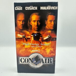 May include: A VHS tape for the movie "Con Air" featuring Nicolas Cage, John Cusack, and John Malkovich. The cover has a fiery background with the film's title and a quote from Siskel & Ebert. The movie title is in a badge with an eagle.