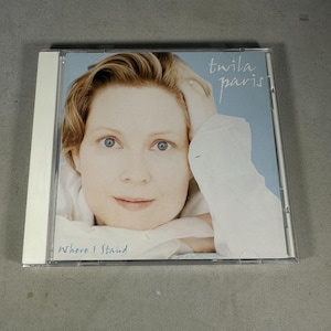 May include: A CD case featuring a woman with short blonde hair and blue eyes. The album cover has a light blue background with the artist's name, "Twila Paris," in cursive. The album title, "Where I Stand," is printed at the bottom.