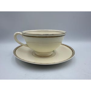 Könnte beinhalten: Cremefarbenes Teetassen- und Untertassen-Set mit goldenem Rand. Die Teetasse hat einen gebogenen Henkel und einen breiten Rand. Die Untertasse ist rund und hat einen passenden goldenen Rand.